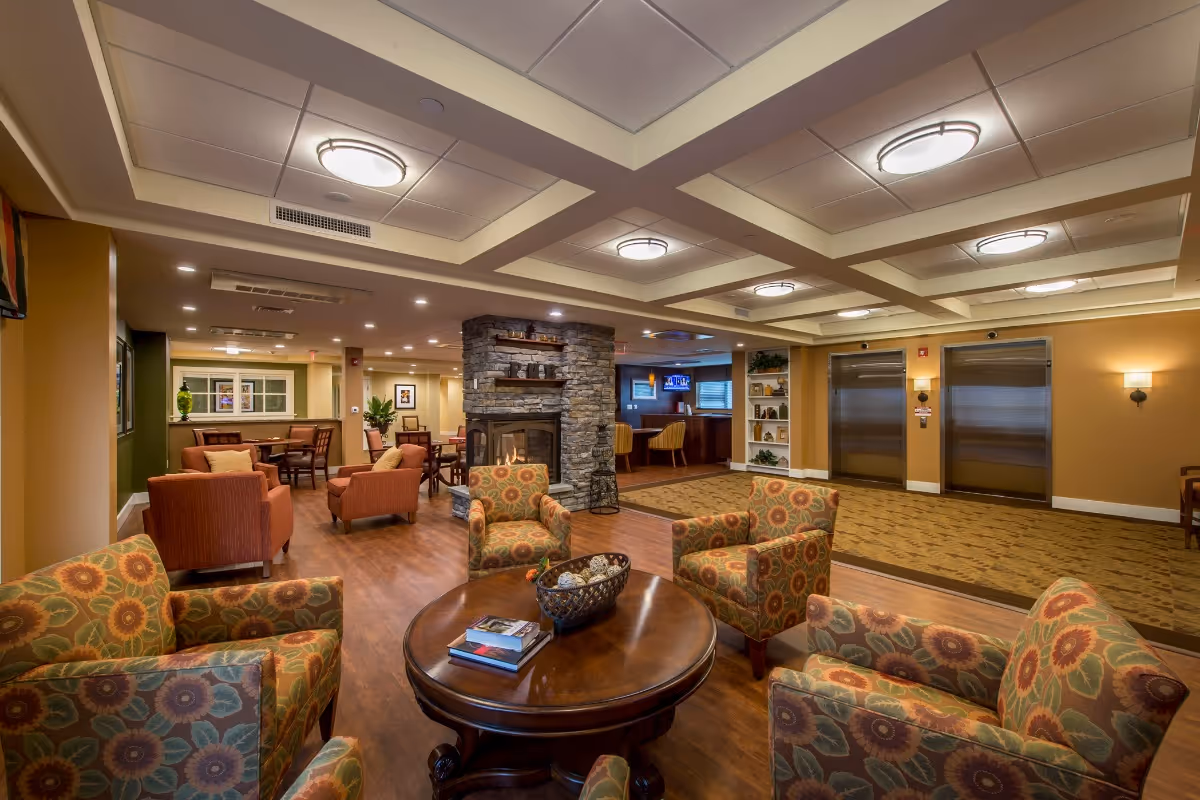 Warm communal lounge with patterned armchairs around a round wooden table, a stone fireplace, and elevators in the background.