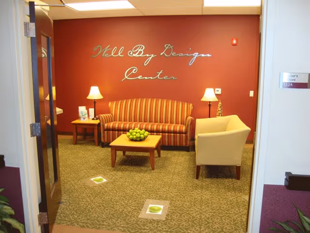 A cozy sitting area with a striped sofa, a beige armchair, two side tables with lamps, and a coffee table with a bowl of green apples. The wall behind the sofa is painted red with the words 'Well By Design Center' written in cursive. The floor is carpeted with a patterned design.