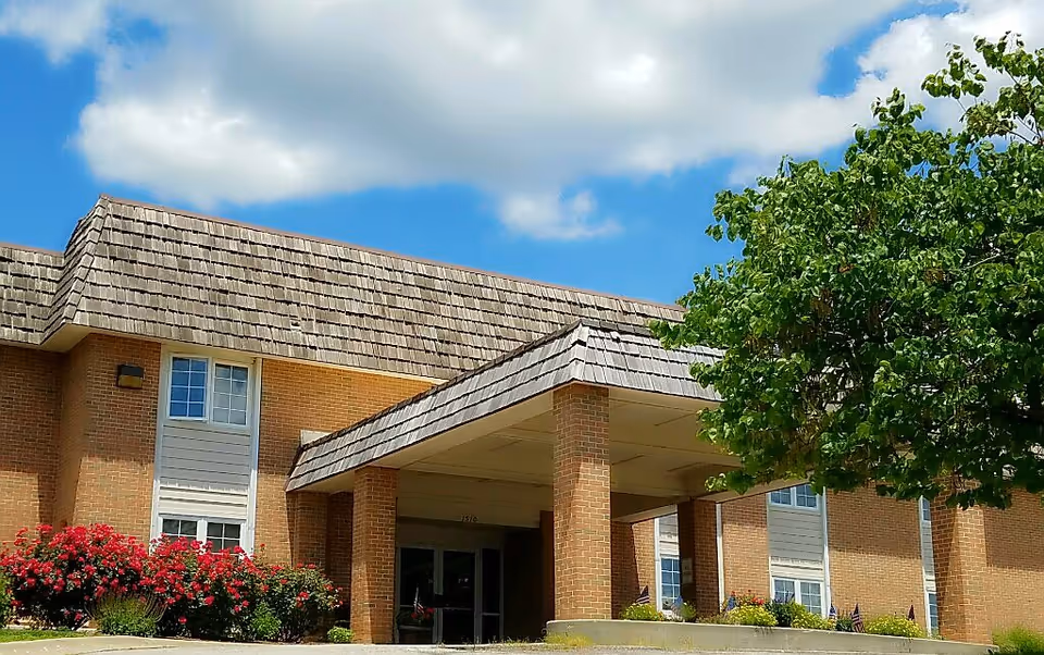 Exterior view of a brick building with a shingled roof and a covered entrance supported by brick columns. There are windows with white frames and flower bushes with red blooms near the entrance. A green tree is visible on the right side under a partly cloudy blue sky.