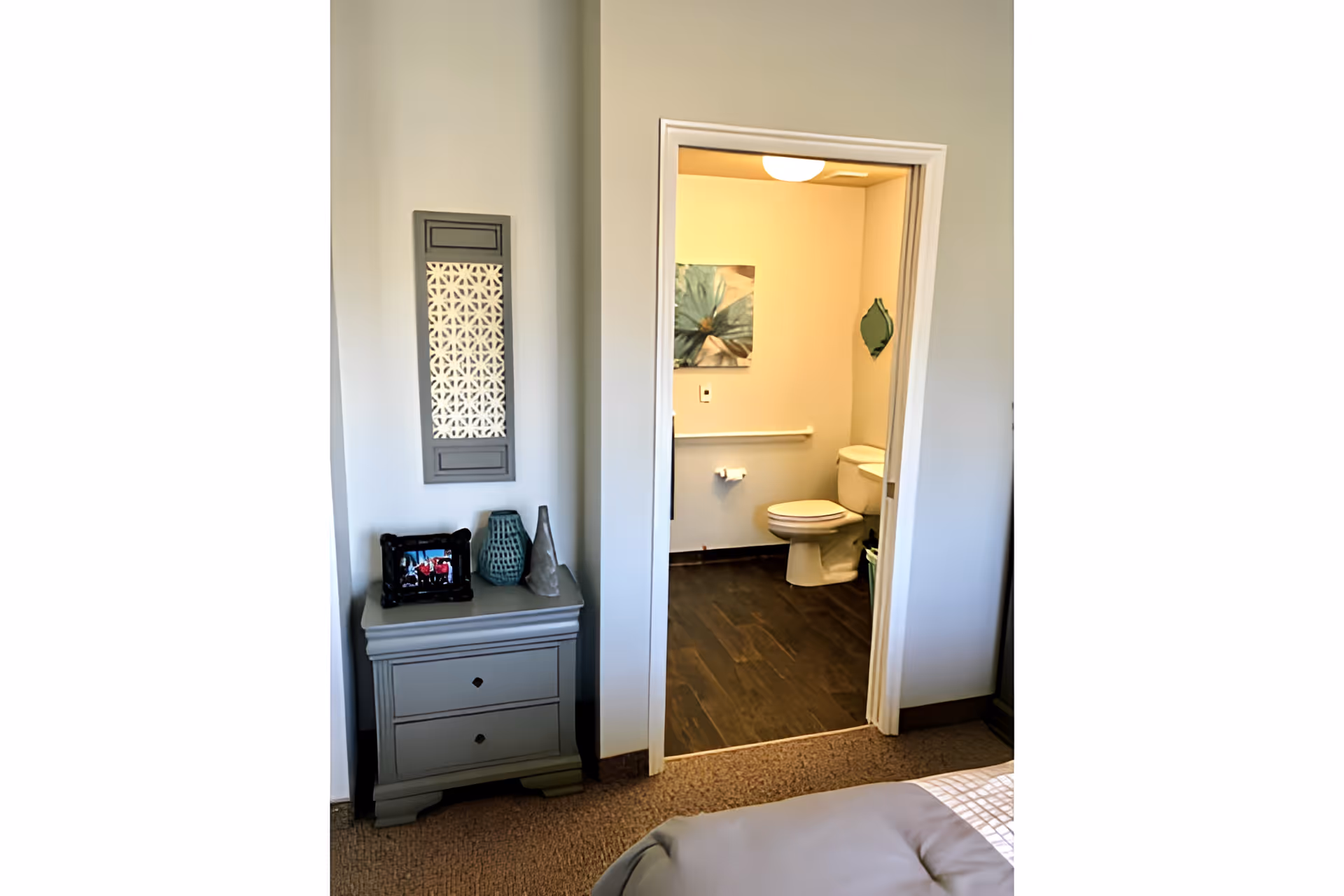 View of a bedroom corner with a gray nightstand holding decorative items and a framed photo, next to an open doorway leading to a bathroom with a toilet, a grab bar, a piece of wall art, and a small green mirror.