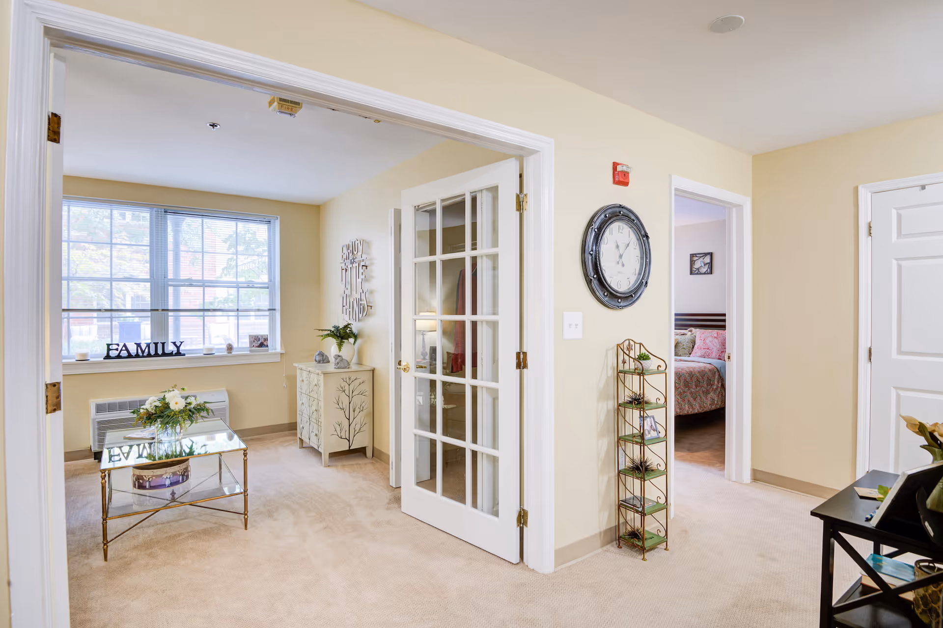 Interior view of a senior living facility showing a bright room with beige walls and carpet. There is a glass coffee table with a flower arrangement, a decorative cabinet, and a window with the word FAMILY displayed on the sill. A large wall clock is mounted on the wall near a doorway leading to a bedroom with a bed and pink bedding. The room has white trim and French doors.