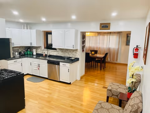 A clean and modern kitchen with white cabinets, black countertops, a stainless steel dishwasher, and a black stove. The kitchen has light wood flooring and recessed ceiling lights. Adjacent to the kitchen is a dining area with a wooden table and four chairs, partially visible through an open doorway. Two patterned chairs with a small table and a yellow flower arrangement are placed against the right wall. A fire extinguisher is mounted on the wall near the dining area.
