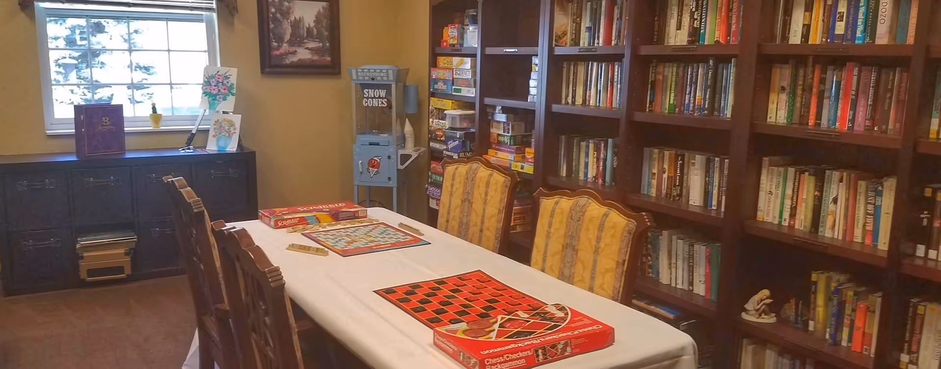 A cozy room with a long table covered with a white tablecloth, set up for board games including Scrabble and Checkers/Backgammon. The room has several wooden chairs with yellow patterned cushions. On the right side, there are tall bookshelves filled with books and board games. A window on the left lets in natural light, and a small cabinet with decorative items is beneath it. A vintage-style snow cone machine is in the corner next to the bookshelves.