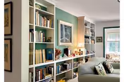 A cozy living room area featuring built-in white bookshelves filled with books and decorative items, a framed artwork hanging on a green accent wall, a table lamp, and a comfortable sofa with patterned cushions near a window with white blinds.