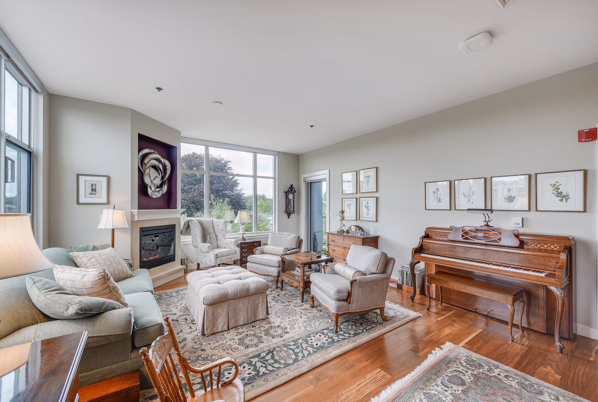 Bright living room with sofas, armchairs, ottoman, a piano, fireplace, and large windows overlooking greenery.