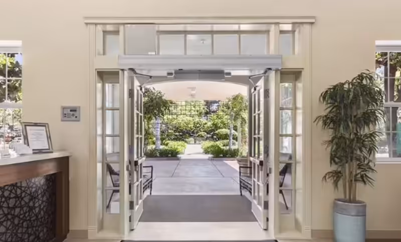 Interior entrance lobby with open double doors opening onto a sunlit courtyard, a reception desk on the left and a potted plant on the right.