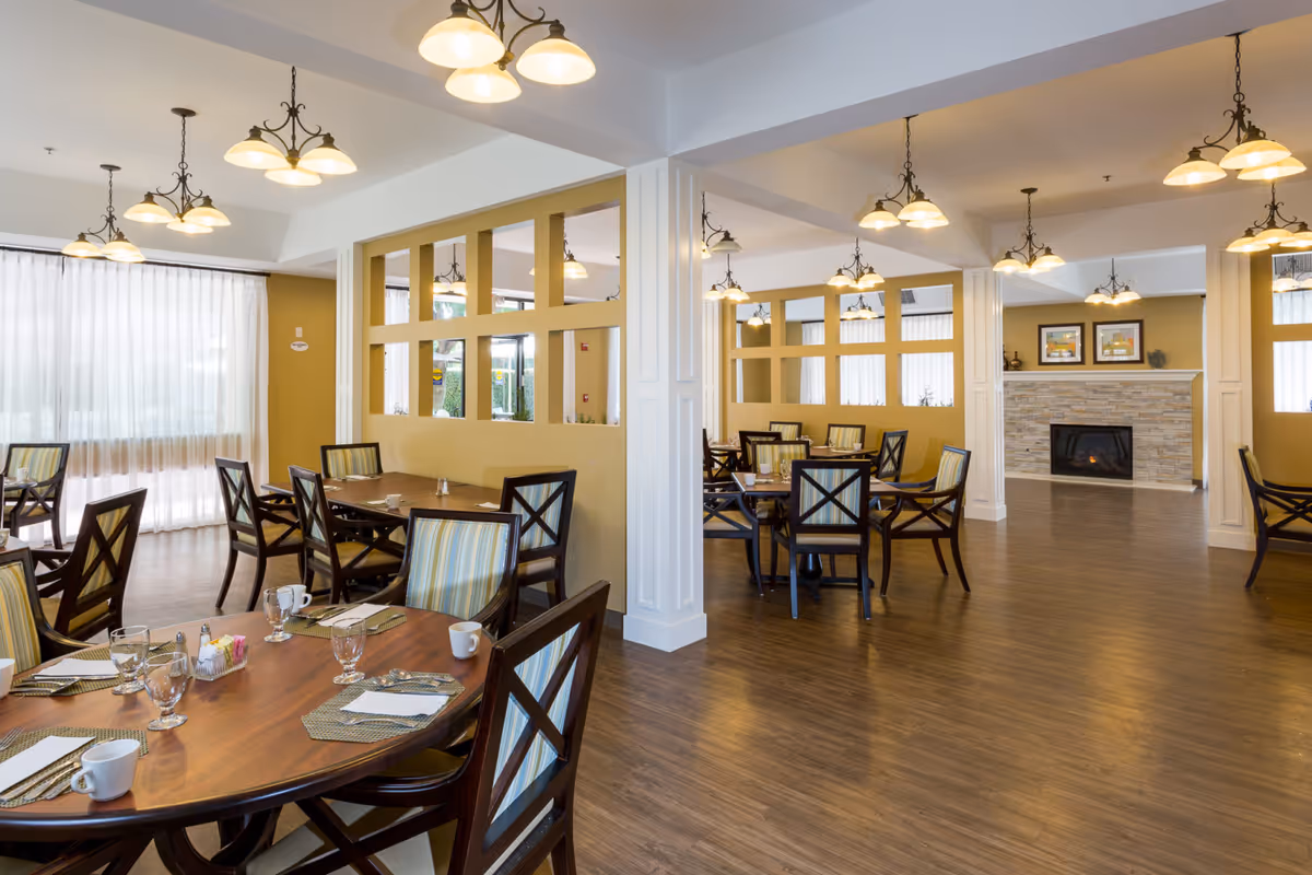 A spacious dining room with multiple wooden tables and chairs, each table set with placemats, cups, glasses, and utensils. The room features warm yellow walls, large windows with sheer white curtains, and several hanging light fixtures. In the background, there is a fireplace with a stone surround and framed artwork above it.