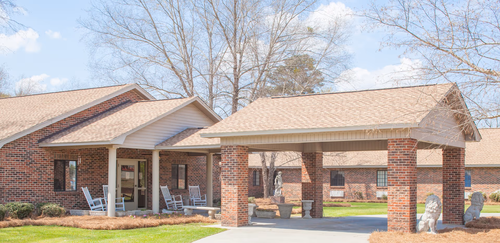 Exterior view of Pembroke Center showing a brick building with a covered entrance supported by brick columns. There are rocking chairs on the porch and decorative lion statues near the driveway. Trees without leaves are visible in the background under a partly cloudy sky.