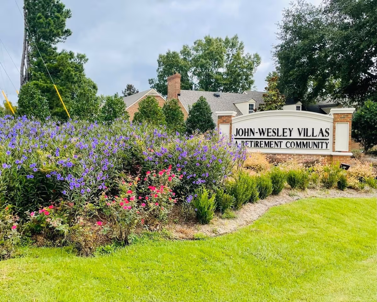 Entrance sign reading 'John-Wesley Villas Retirement Community' set beside flowering shrubs and a manicured lawn with buildings and trees in the background.