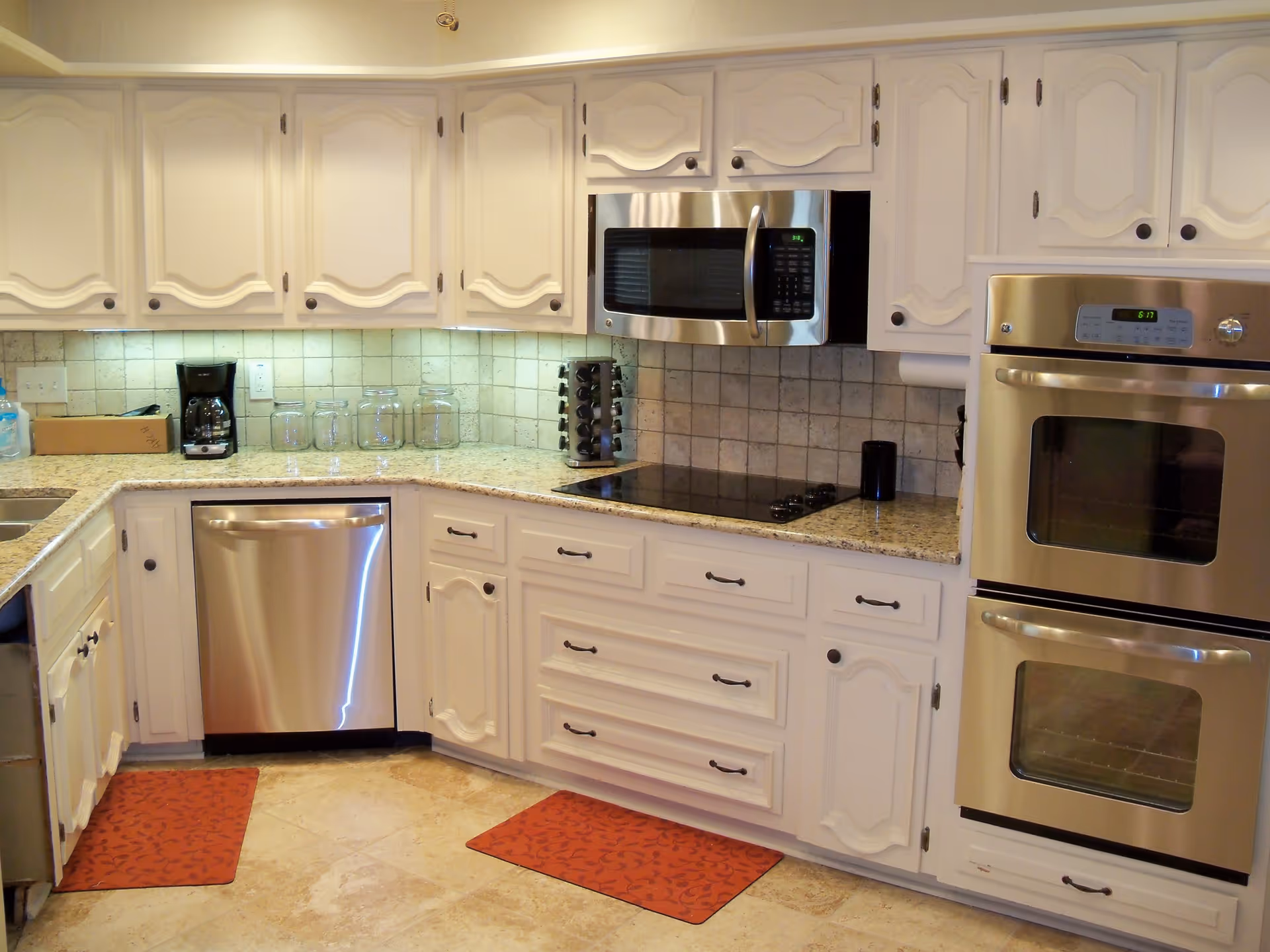 A clean and modern kitchen with white cabinets, granite countertops, and stainless steel appliances including a dishwasher, microwave, and double oven. There are two red patterned floor mats, a coffee maker, glass jars, and a spice rack on the countertop. The backsplash is tiled in a beige color.