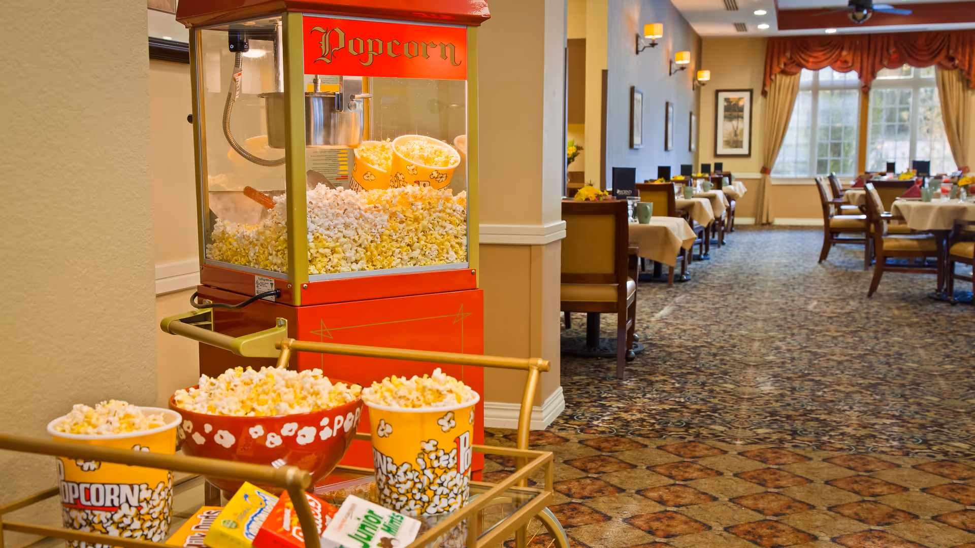A popcorn machine filled with freshly popped popcorn next to a cart holding three bowls of popcorn in a dining area with tables and chairs set up for meals, featuring carpeted flooring and large windows with curtains in the background.