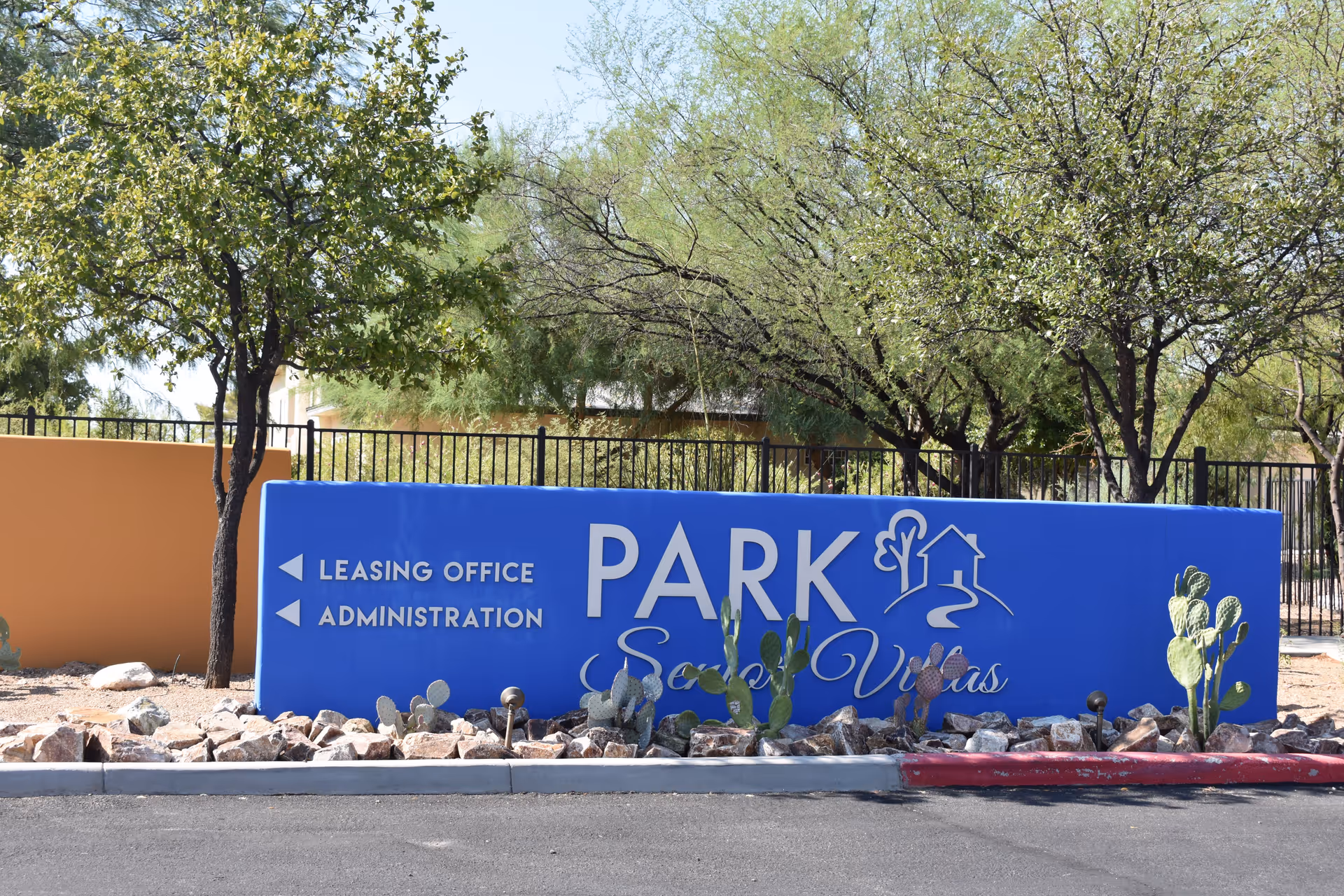 Outdoor view of a blue sign for Park Senior Villas with arrows pointing left for Leasing Office and Administration, surrounded by desert landscaping with rocks, cacti, and trees.