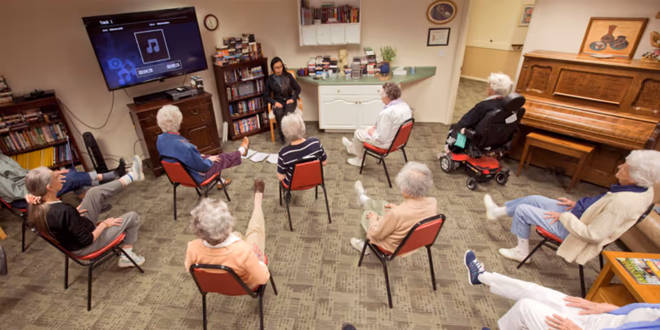 Elderly residents seated in a community room raise their legs during a seated exercise class led by a staff member.