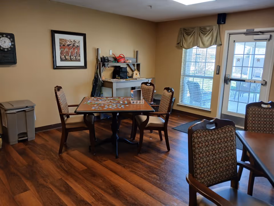 Communal dining/activity room with a table and chairs, a jigsaw puzzle on the table, shelving, window and a door to the outside.