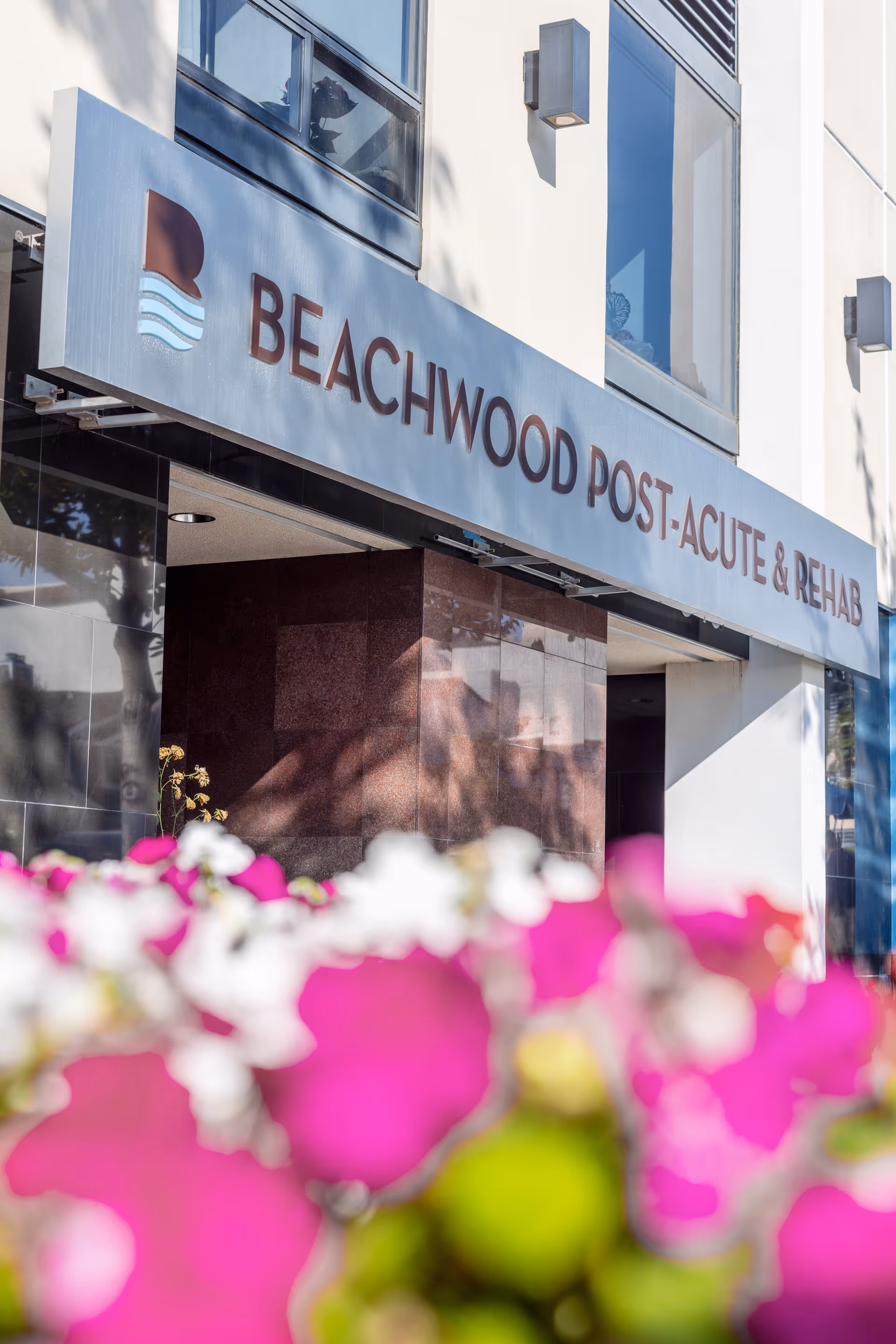 Entrance of Beachwood Post-Acute & Rehab facility with a sign above the doorway and pink and white flowers in the foreground.