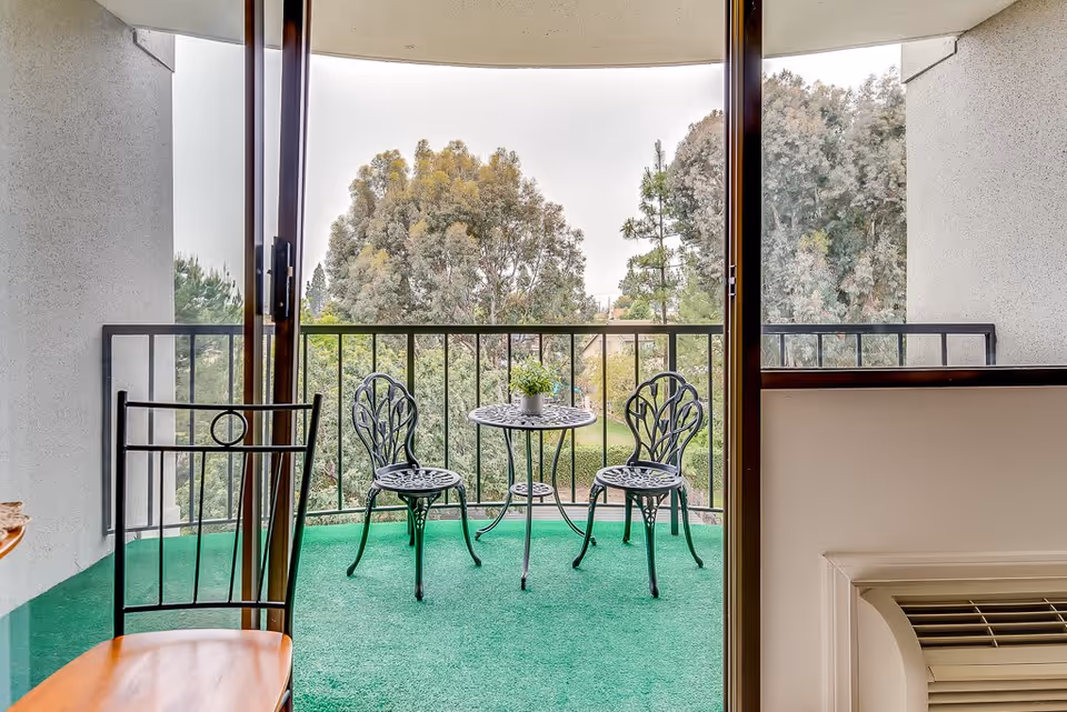 View through a sliding glass door onto a balcony with green carpet, featuring a small round metal table with a potted plant and two matching metal chairs. Trees and greenery are visible beyond the balcony railing.