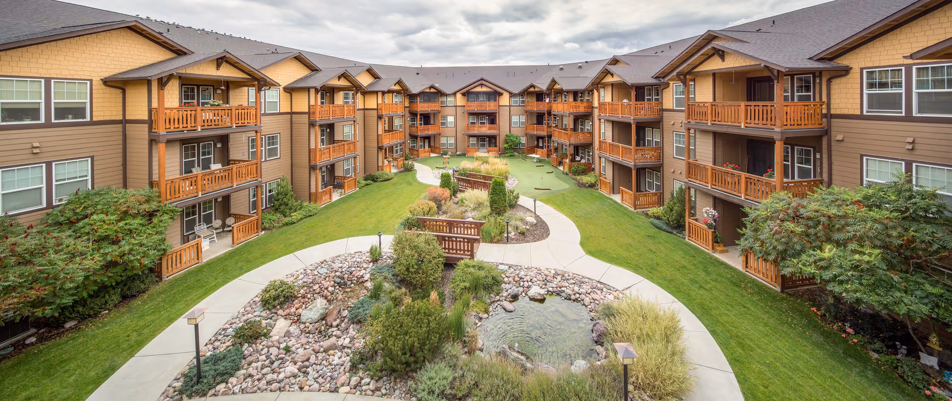 View of a senior living facility courtyard with a landscaped garden, walking paths, a small pond, and wooden benches. The surrounding buildings have multiple floors with balconies and large windows, featuring a brown and tan exterior with wooden railings.