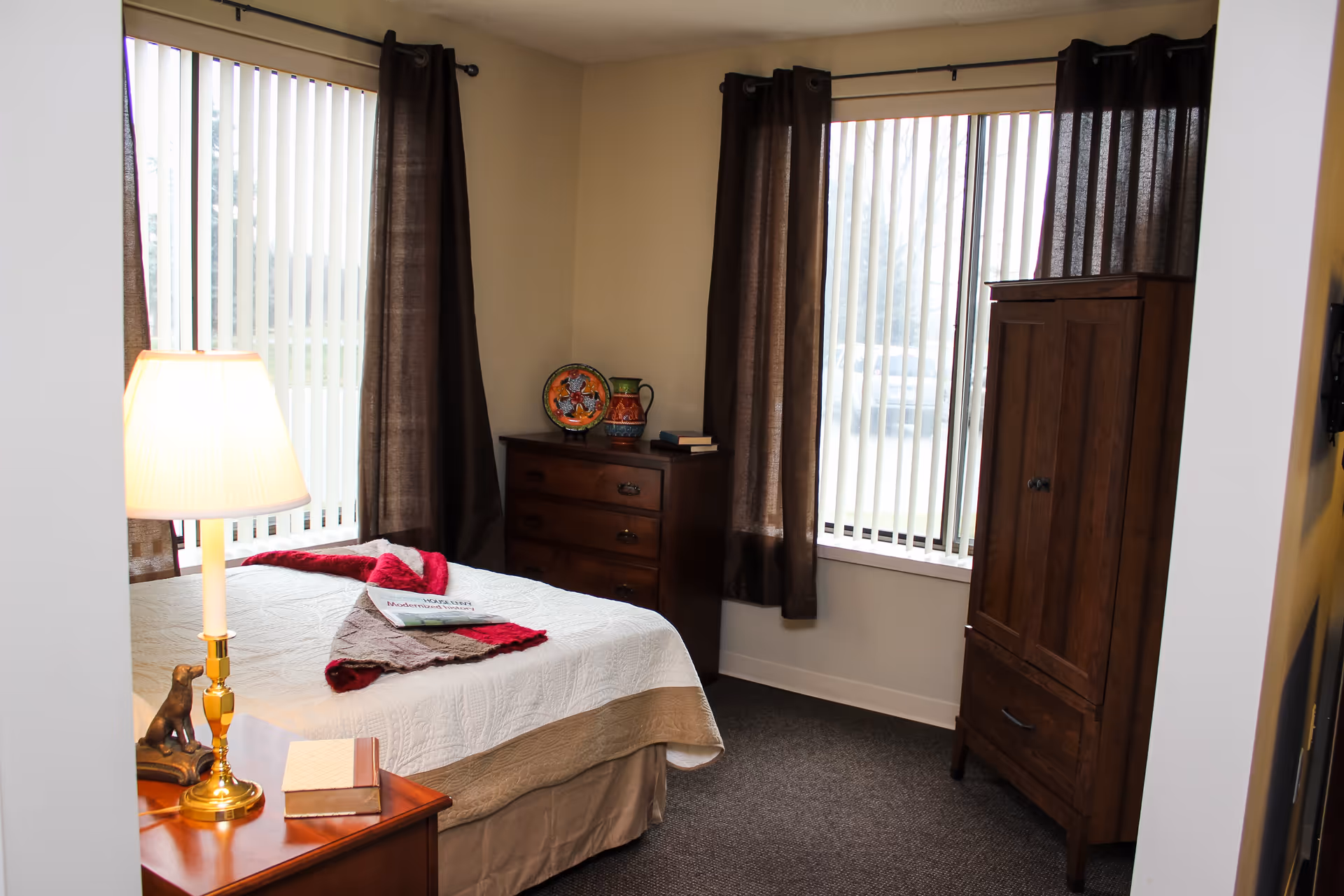A cozy bedroom with a bed covered in a white quilt and a red and brown throw blanket. There is a wooden nightstand with a lamp and a book, a wooden dresser with decorative items and books, and a wooden wardrobe. The room has two large windows with vertical blinds and dark brown curtains.