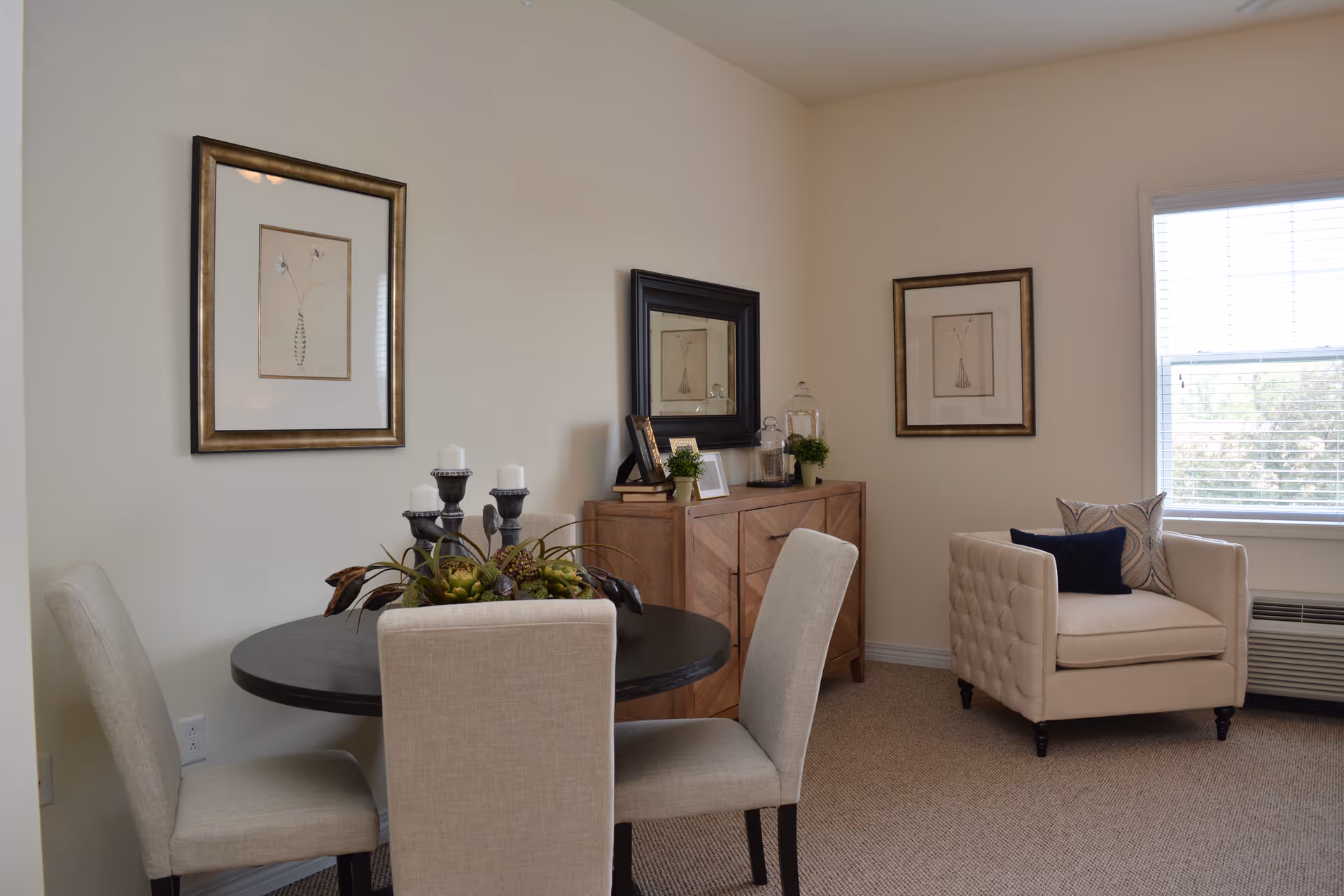 A cozy interior room featuring a round black dining table with four beige upholstered chairs. On the table is a decorative centerpiece with candles and greenery. Against the wall is a wooden sideboard with framed pictures, plants, and decorative jars. Two framed botanical prints hang on the walls, and a beige armchair with two pillows sits near a window with blinds, allowing natural light to fill the room.