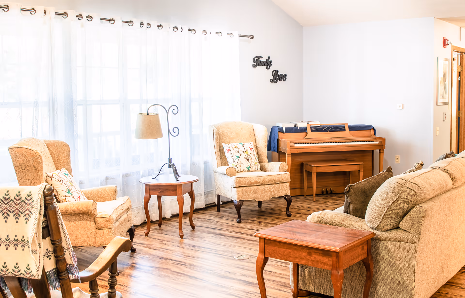Bright living room with upholstered armchairs and a sofa, wooden side tables, a floor lamp, and an upright piano by a window with sheer curtains.