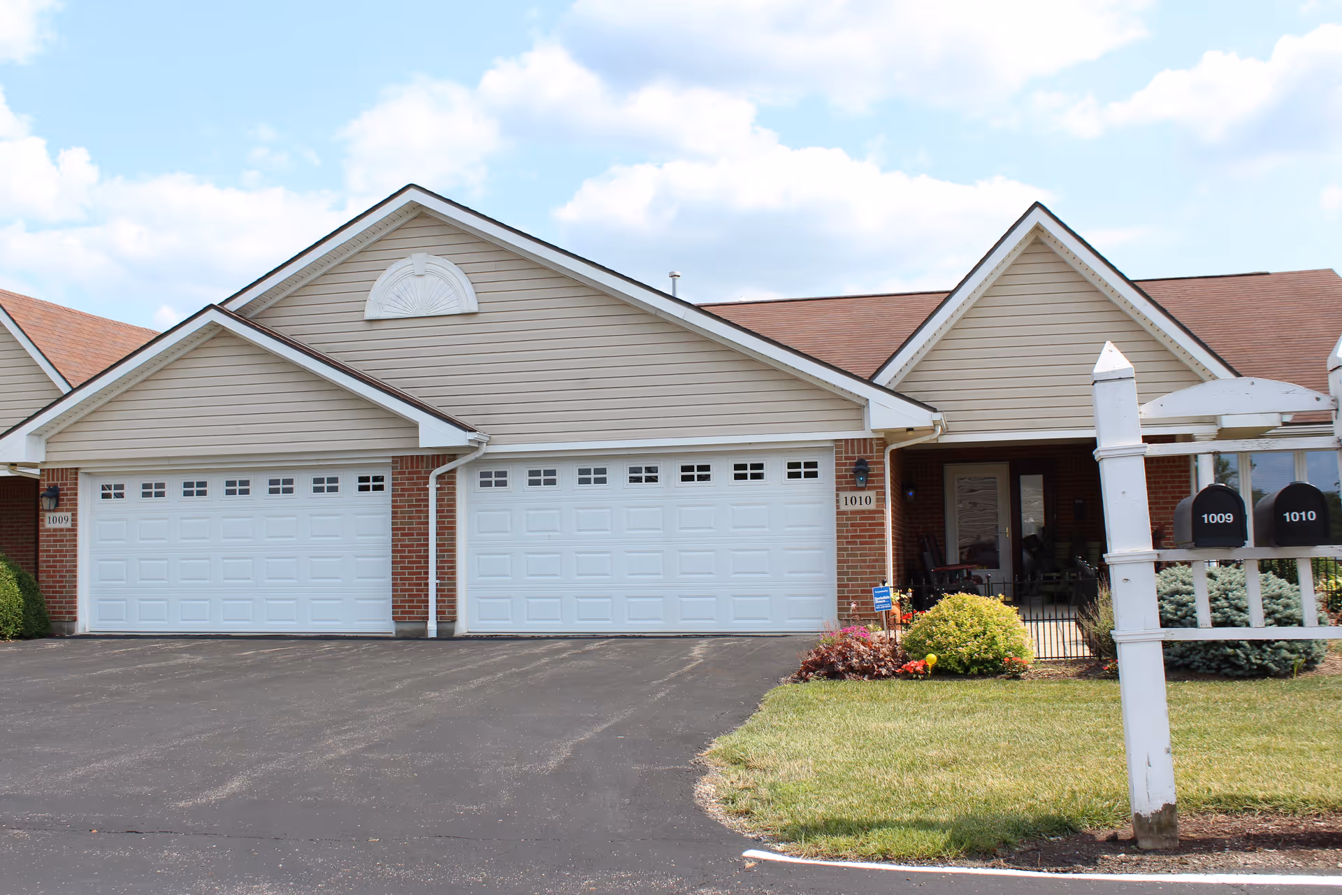 Exterior view of a residential building at Brookhaven Retirement Community featuring two attached garages with white doors, beige siding, and brick accents. The building has a well-maintained lawn with shrubs and a white mailbox post displaying numbers 1009 and 1010. The sky is partly cloudy.