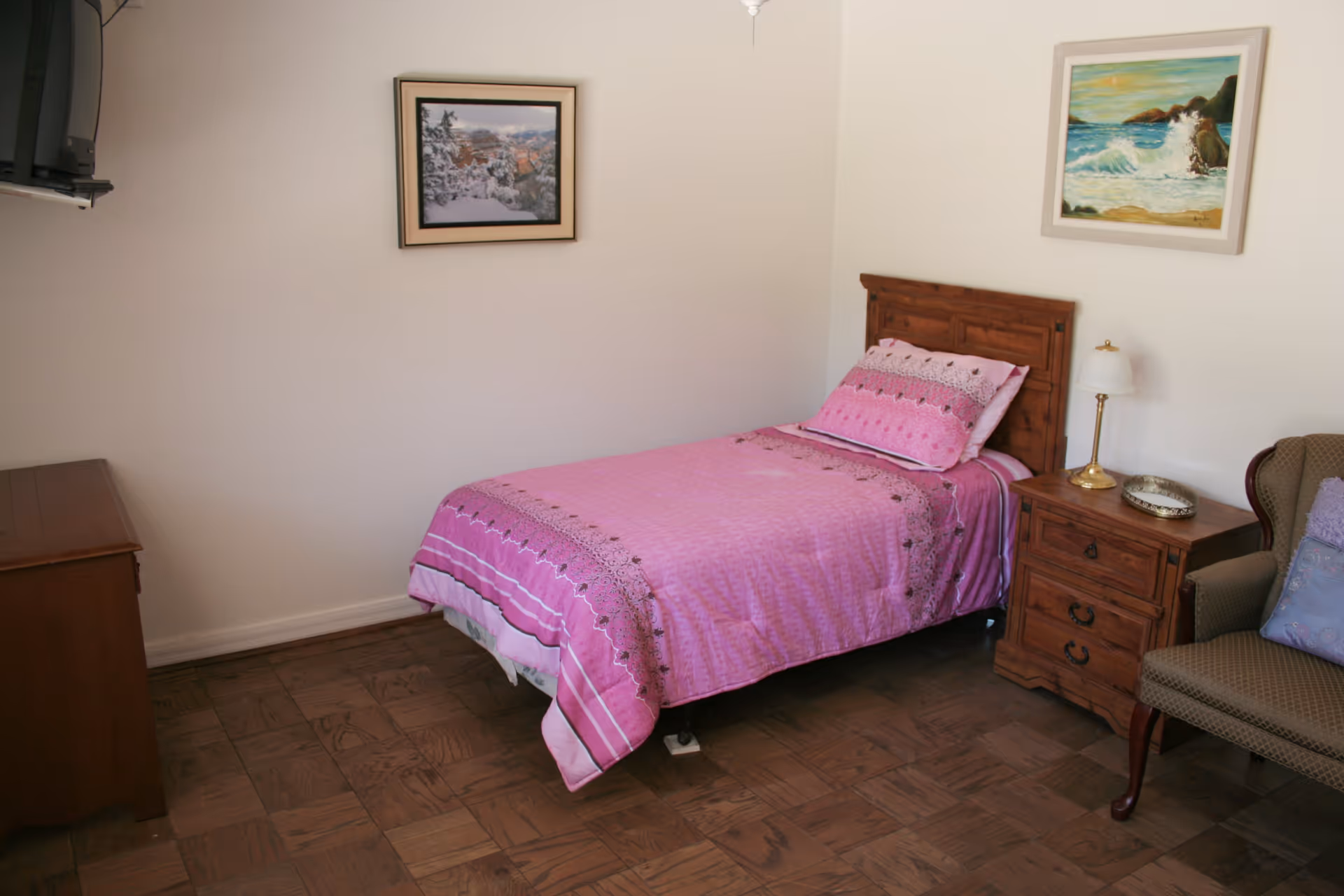 A simple bedroom with a single bed covered in a pink bedspread. There is a wooden nightstand with a lamp and a decorative plate next to the bed. A cushioned armchair with a pillow is positioned beside the nightstand. Two framed paintings hang on the walls, one above the bed and one on the adjacent wall. The floor is wooden parquet, and a wooden dresser is partially visible on the left side of the image.
