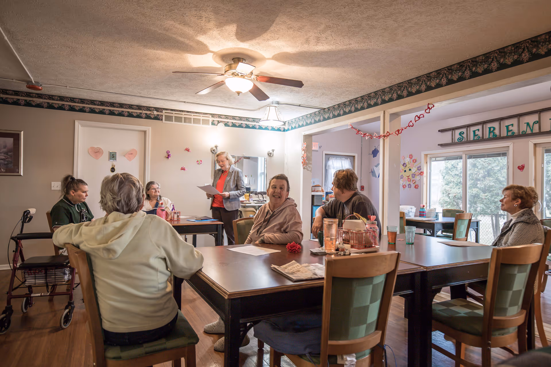 A group of elderly residents sit around a dining table in a decorated common room while a staff member stands reading papers.
