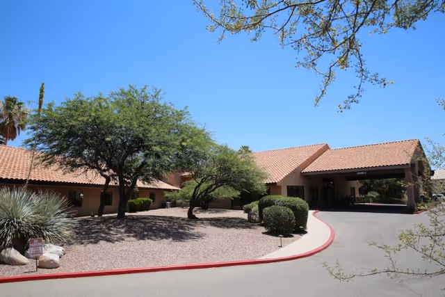 Exterior view of La Cañada Care Center showing a single-story building with a tiled roof, surrounded by desert landscaping including trees, bushes, and gravel. The driveway curves around the front entrance under a covered drop-off area.