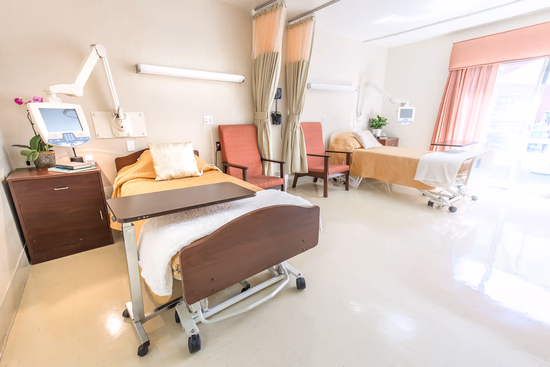 A bright and clean hospital or healthcare facility room with two beds separated by a curtain. Each bed has a bedside table with medical equipment and a plant. There are two red chairs between the beds and large windows with pink curtains allowing natural light to fill the room.