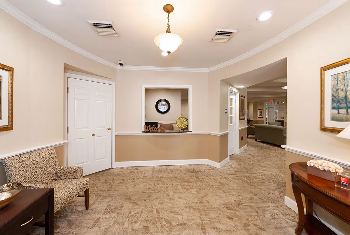 A clean and well-lit reception area in a senior living facility with beige walls and carpeted floor. There is a small reception window with a 'Welcome' sign and decorative items. To the left, there is a patterned armchair next to a small table with a decorative bowl. On the right, a wooden console table holds a lamp and some decorative items. The hallway leads to a lounge area with sofas and framed artwork on the walls.