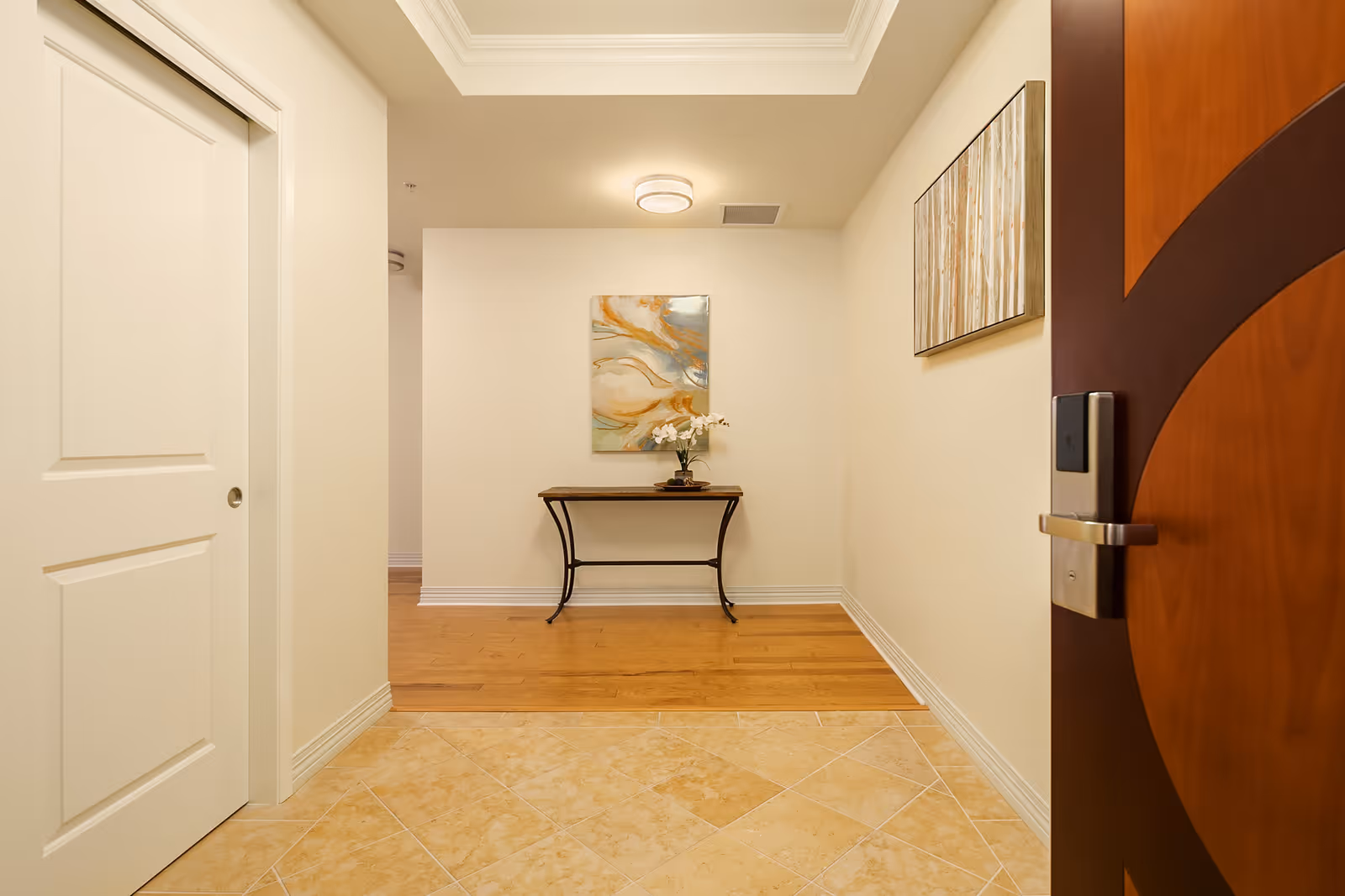 View of an entryway in a senior living facility with a wooden door partially open on the right, beige tiled floor transitioning to wooden flooring, a small table with a flower vase against the far wall, and two abstract paintings hanging on the walls.