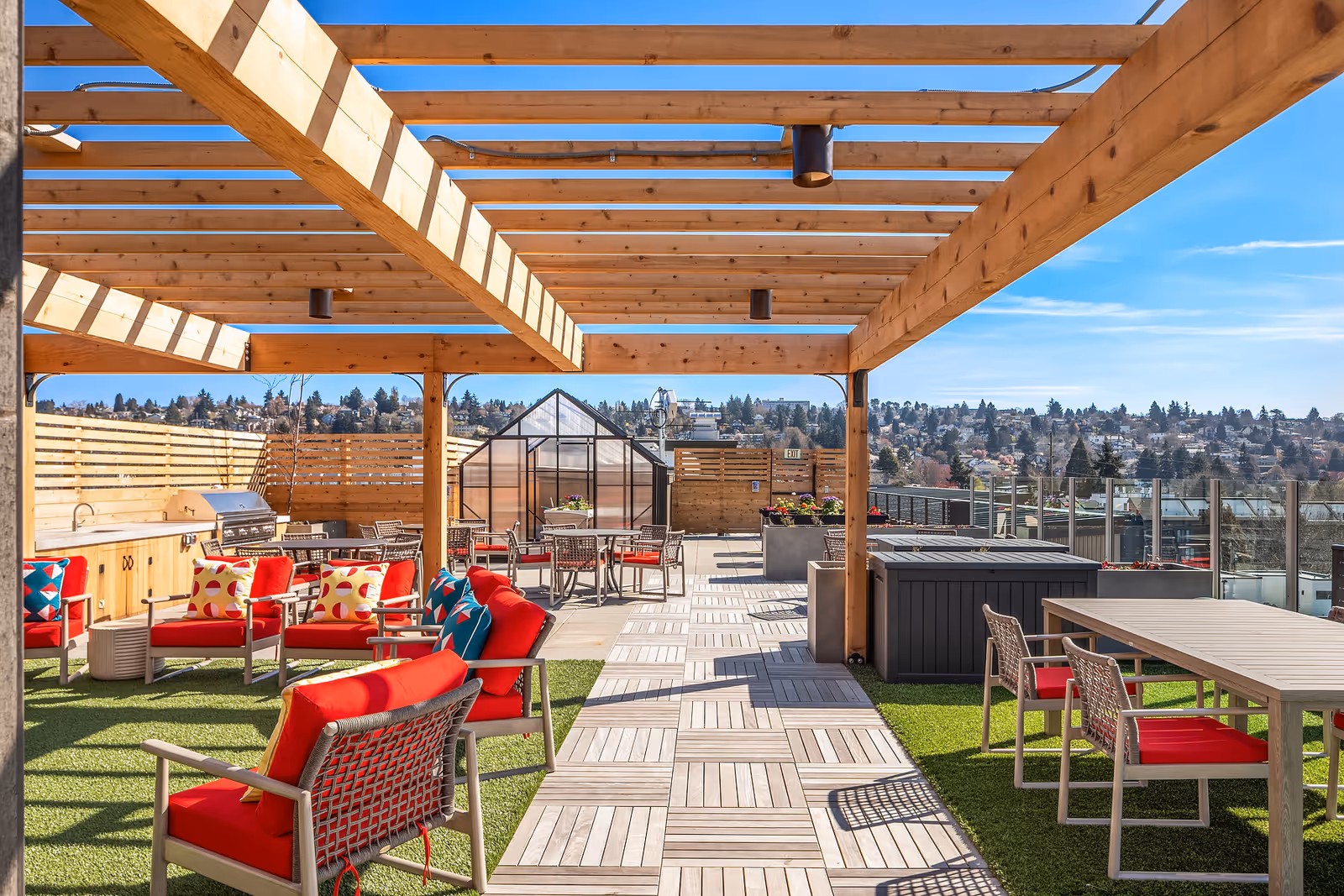 Outdoor rooftop patio area with wooden pergola overhead, red cushioned chairs and sofas, tables with chairs, a barbecue grill, and a small greenhouse in the background. The area overlooks a cityscape under a clear blue sky.