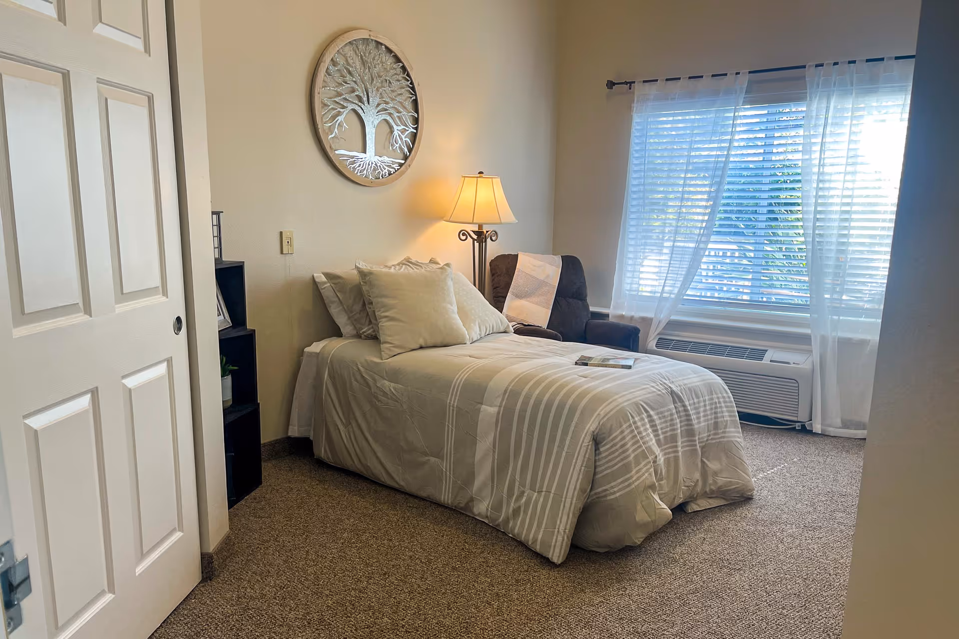 A cozy bedroom with a single bed covered in beige and white striped bedding, two pillows, and a book placed on top. Next to the bed is a dark brown armchair with a white throw blanket draped over it. A floor lamp with a beige shade is positioned between the bed and the chair. A large window with white blinds and sheer curtains allows natural light to fill the room. On the wall above the bed hangs a round decorative piece featuring a tree design. The floor is carpeted in a neutral tone, and a white door is partially open on the left side of the image.