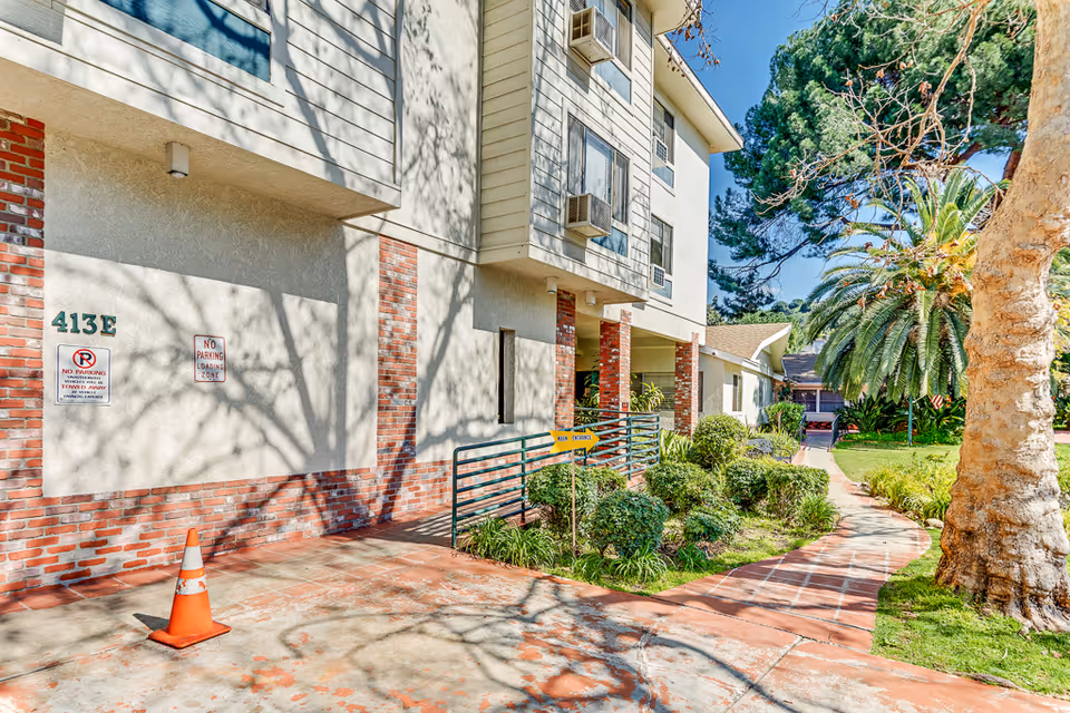 Exterior view of a building with brick and light-colored siding, featuring multiple windows with air conditioning units. A sidewalk curves through a landscaped area with bushes, a large tree, and a palm tree. There are signs indicating no parking and a loading zone, as well as a small yellow sign pointing to the main entrance.
