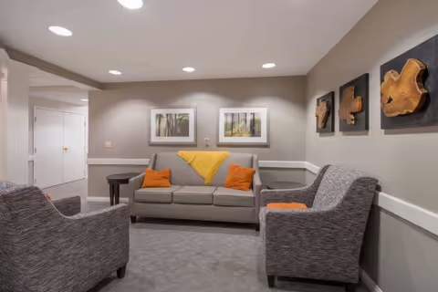 A cozy sitting area with a beige sofa adorned with orange pillows and a yellow throw blanket, flanked by two gray armchairs with orange cushions. The walls are decorated with two framed landscape pictures and three wooden art pieces. The room has recessed ceiling lights and neutral-colored walls and carpet.