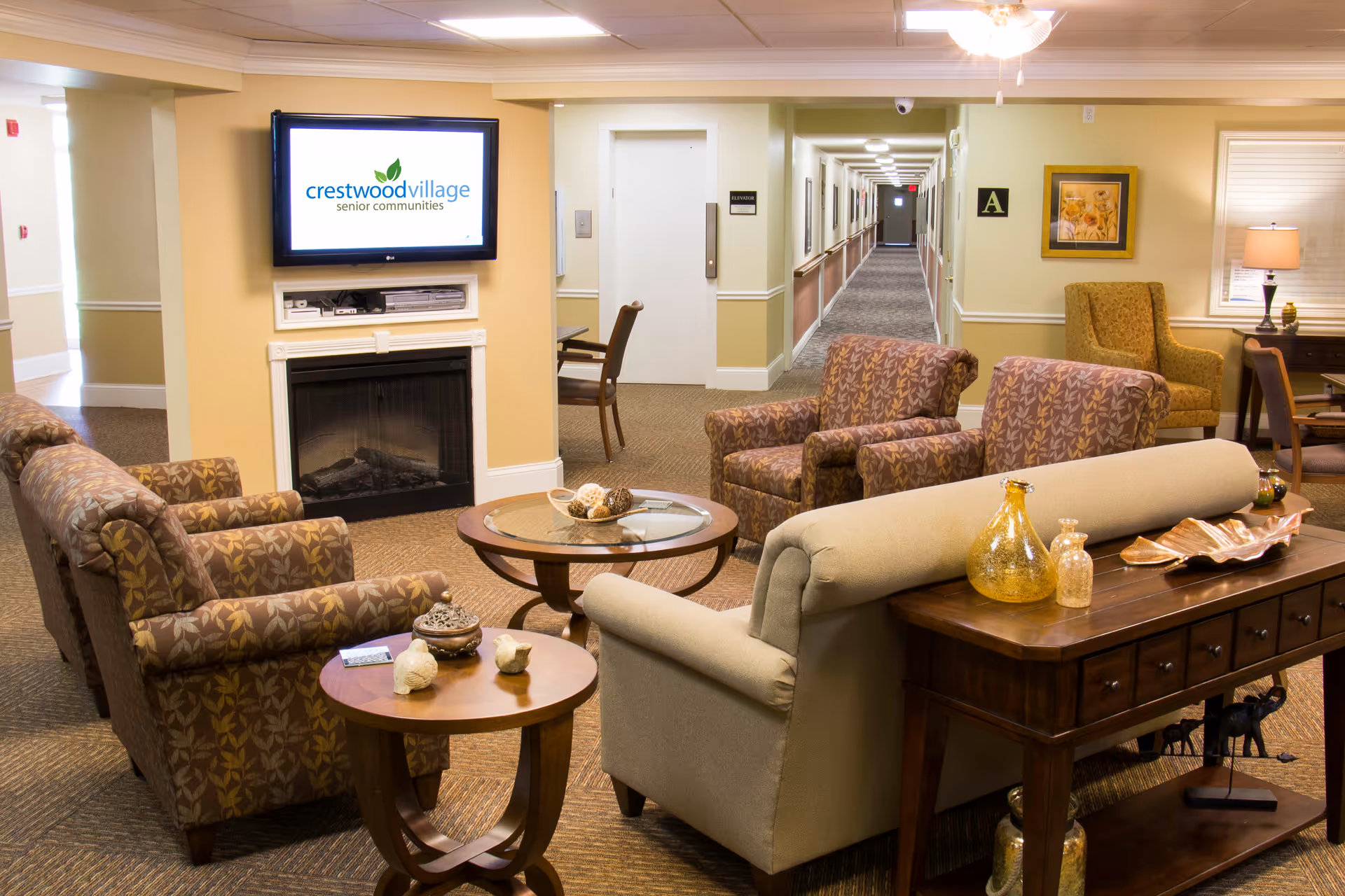 A cozy senior living community lounge area with patterned armchairs and a beige sofa arranged around a glass coffee table and a side table with decorative items. A flat-screen TV mounted above a fireplace displays the Crestwood Village Senior Communities logo. The background shows a long hallway with multiple doors and a chair near an elevator.