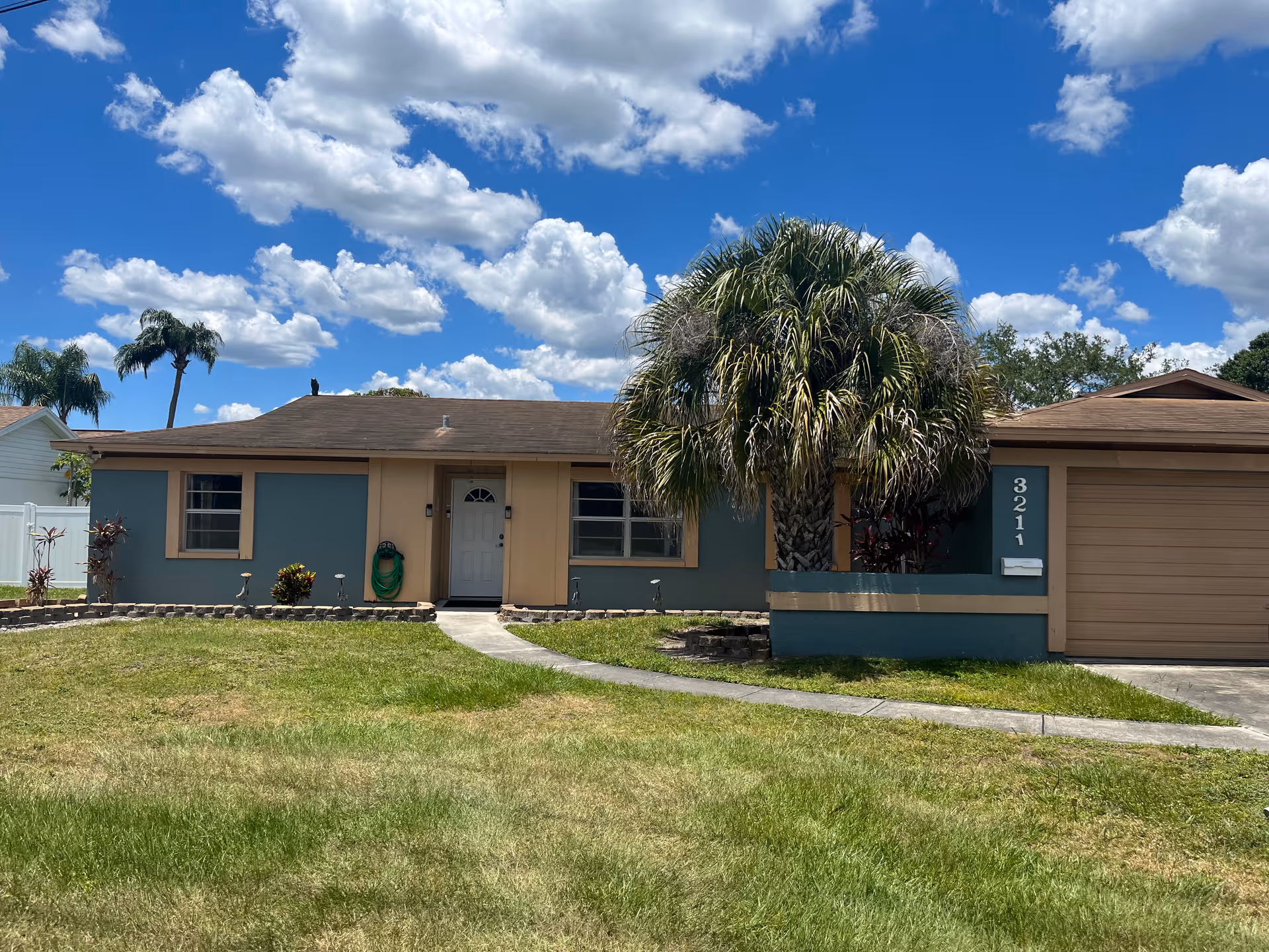 Single-story residential building with a brown roof and teal walls with beige trim. There is a white front door in the center, two windows on either side, and a garage on the right. A large palm tree is in front of the garage area. The house number 3211 is displayed vertically near the garage. The lawn is green with a curved concrete walkway leading to the front door. The sky is bright blue with scattered white clouds.