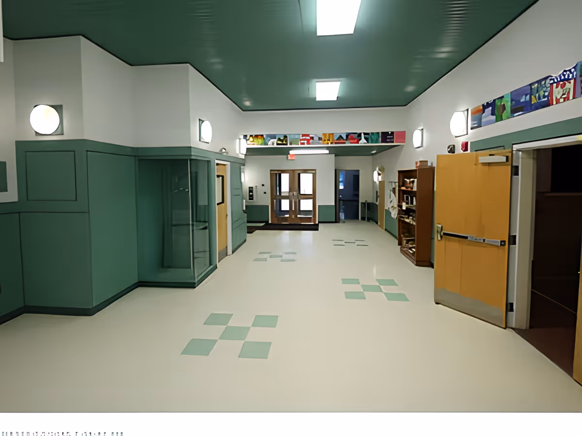 Interior hallway of Frankfort Assisted Living facility with green and white walls and ceiling, light fixtures on the walls, a display case on the left, wooden double doors at the far end, and an open wooden door on the right side.