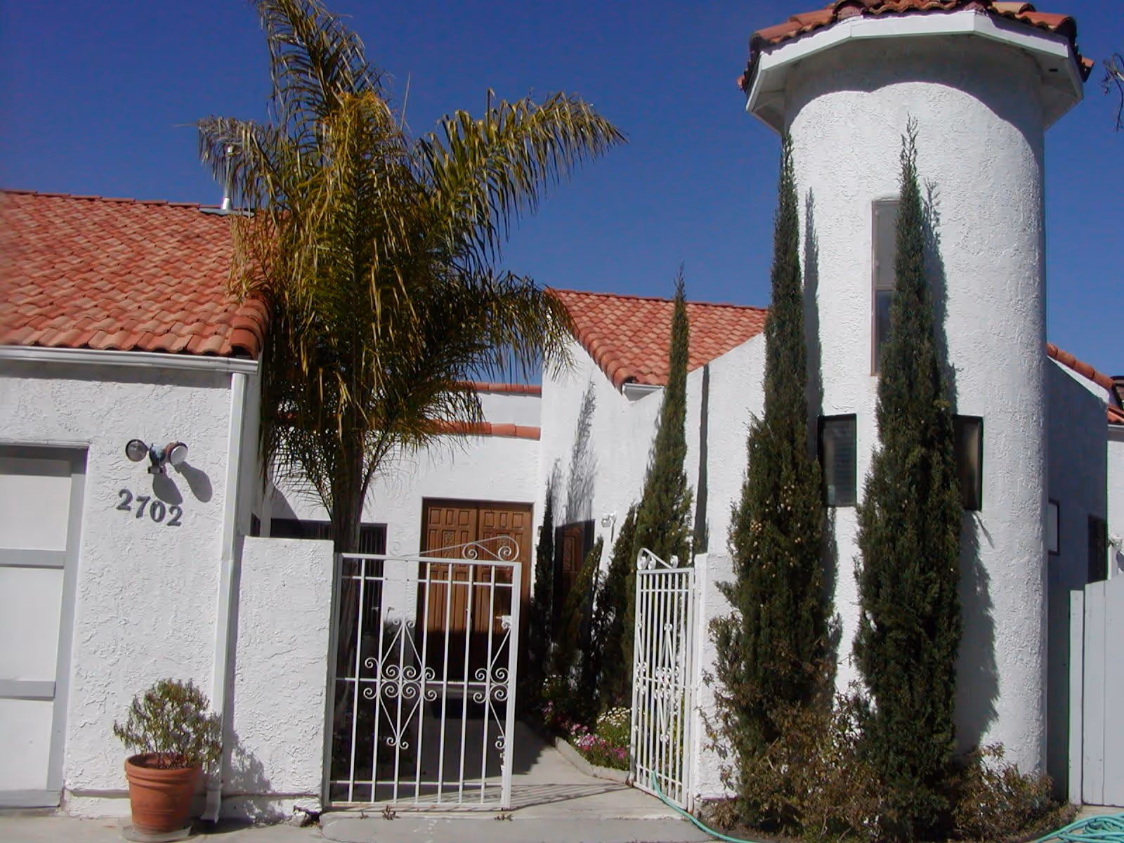 Exterior view of a white stucco building with a red tile roof, featuring a round tower-like structure and tall narrow windows. There is a white metal gate at the entrance, several tall cypress trees, a palm tree, and a potted plant near the garage door with the number 2702. The sky is clear and blue.