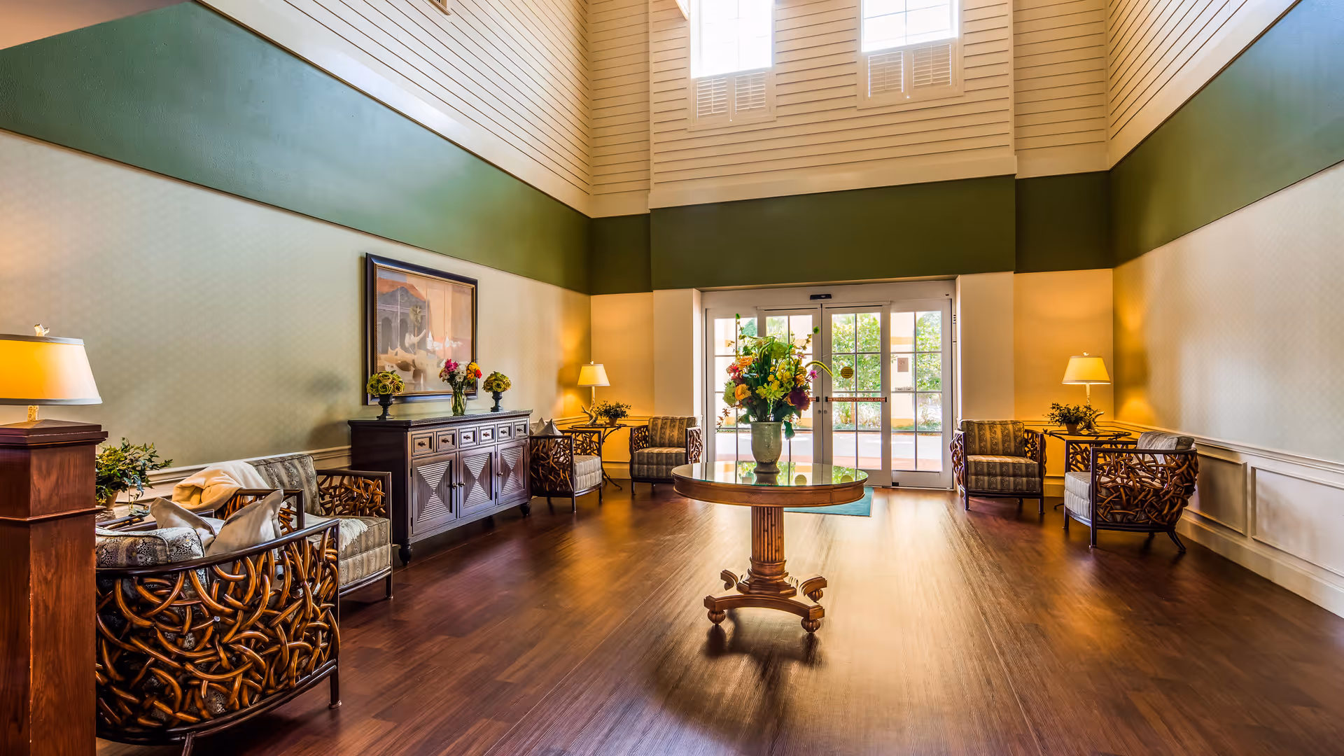 A spacious and well-lit living room area with high ceilings and large windows. The room features a round wooden table with a flower arrangement in the center, several cushioned armchairs with wooden frames arranged along the walls, two side tables with lamps, a wooden cabinet with decorative items and flowers, and a glass door leading outside.
