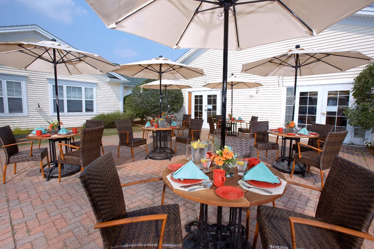 Outdoor patio area with multiple round tables set for dining, each table has brown wicker chairs, white umbrellas, red plates, red mugs, blue napkins, and flower centerpieces. The patio is paved with bricks and surrounded by white buildings with windows and doors.