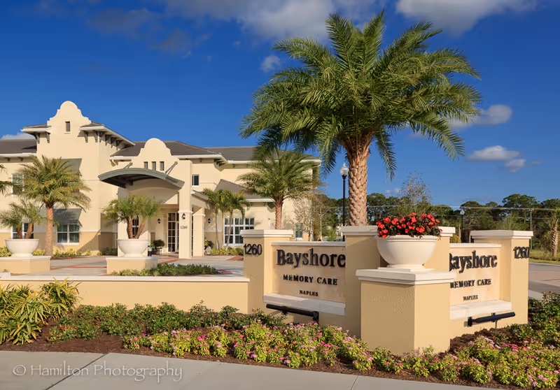 Exterior view of Bayshore Memory Care facility in Naples, featuring a beige building with palm trees and landscaped flower beds under a blue sky with some clouds. The facility's name and address, 1260, are displayed on a sign in front.