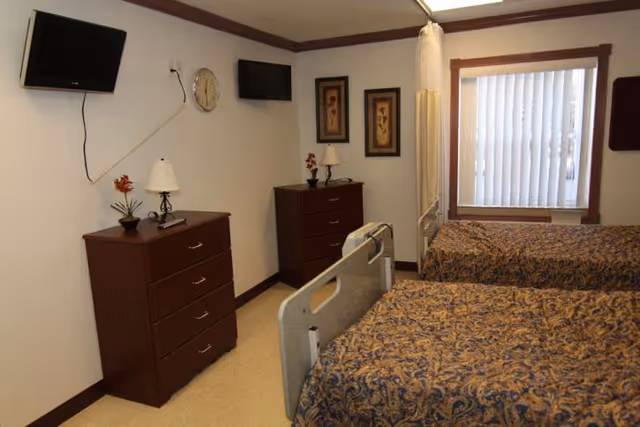 Two hospital-style beds with patterned bedspreads, bedside dressers, wall-mounted TVs and a window with vertical blinds in a senior care room.