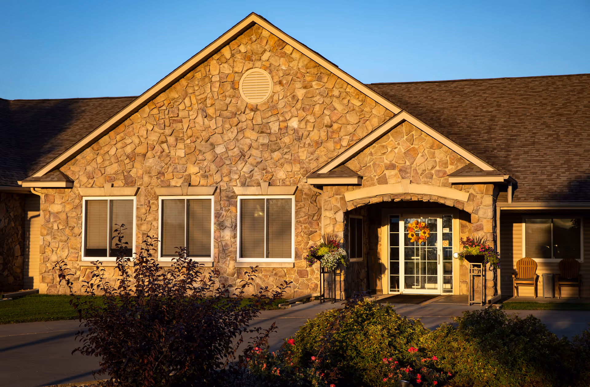 Exterior view of a stone-faced building with three windows and a glass door entrance decorated with a sunflower wreath. There are flower pots on either side of the entrance and two wooden chairs to the right. The building is illuminated by warm sunlight and surrounded by bushes and greenery.