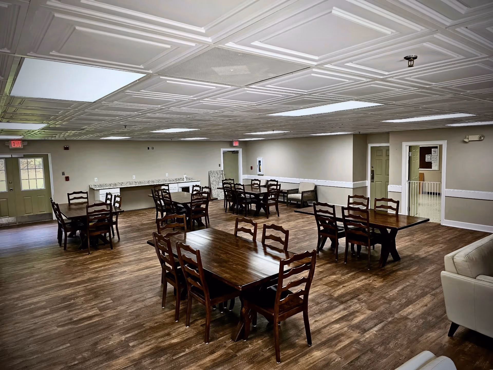 A spacious dining room with wooden tables and chairs, featuring a decorative ceiling and large windows. The room has a warm, inviting atmosphere with a light-colored wall and a counter area in the background.