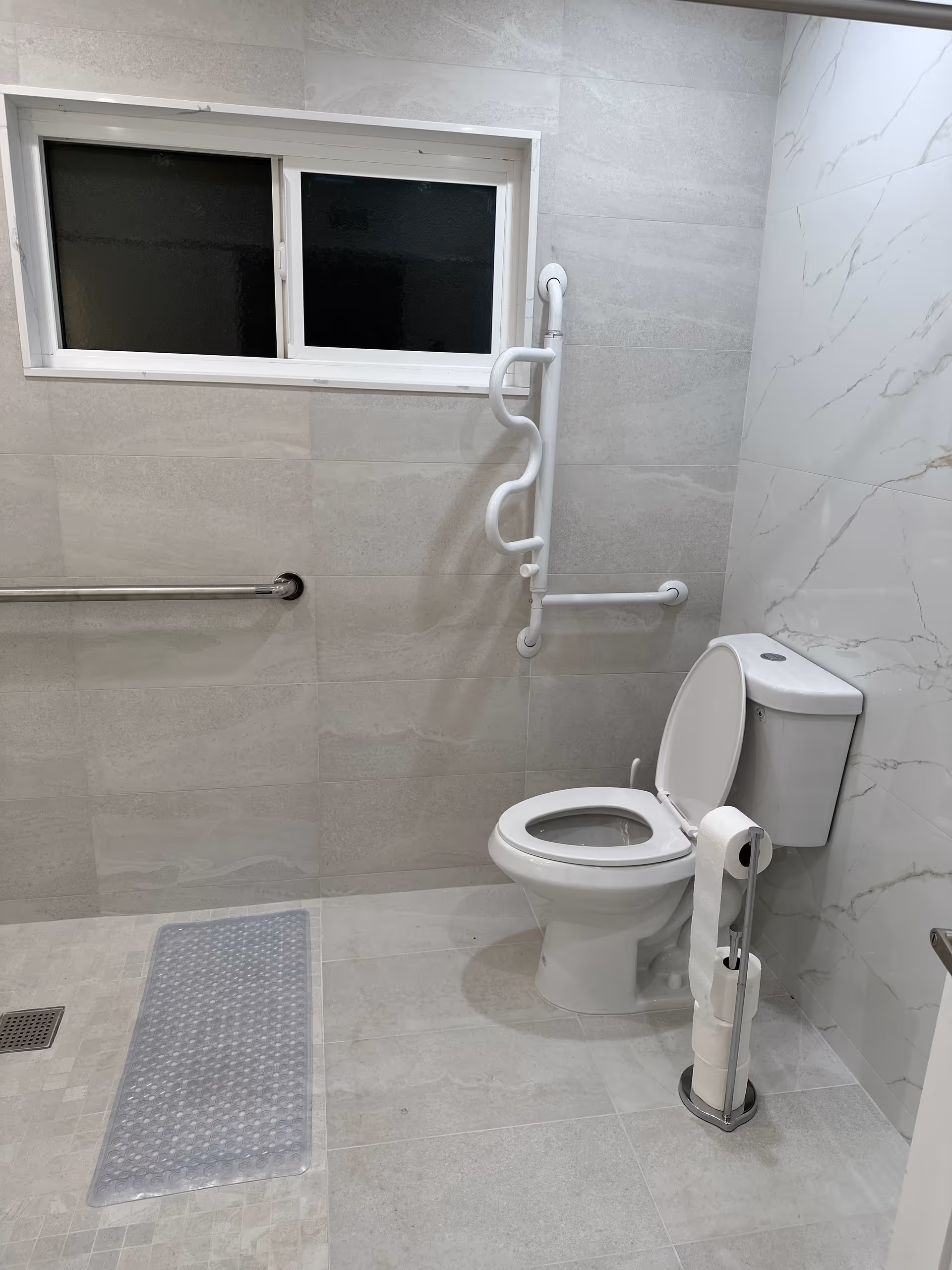 A clean, accessible bathroom with a white toilet, grab bars on the wall for support, a frosted window, and a toilet paper holder with multiple rolls. The walls and floor are tiled in light gray tones.