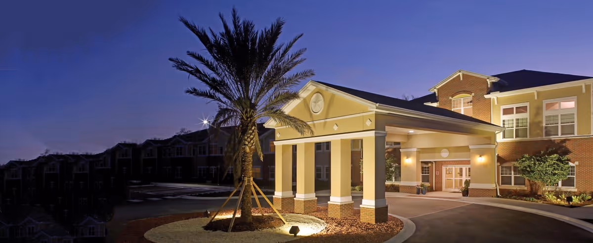 Exterior view of Anthem Lakes senior living facility at dusk, showing the entrance with a covered driveway, a tall palm tree in front, and the building illuminated by exterior lights.