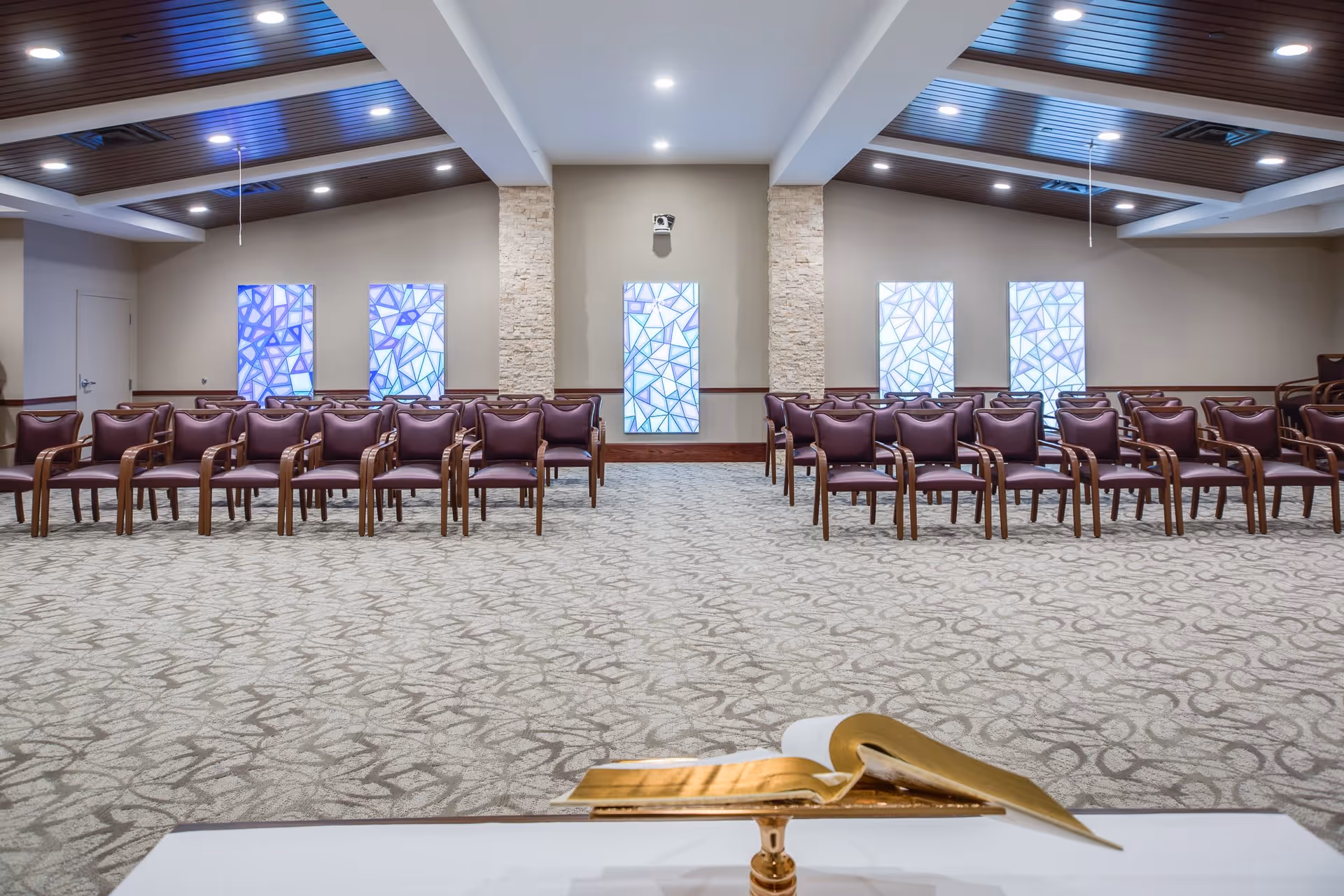 Interior view of a large room with rows of brown chairs arranged facing a podium with an open book. The room has a patterned carpet, beige walls, stone columns, and decorative stained glass panels on the far wall under a ceiling with recessed lighting.