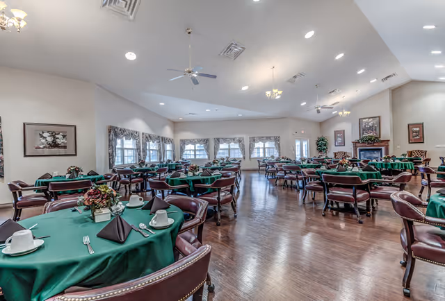 A spacious dining room with multiple round tables covered in green tablecloths, each set with white cups, saucers, silverware, and black napkins. The room has wooden floors, several windows with floral curtains, ceiling fans, and framed artwork on the walls. There is a fireplace with plants and chairs arranged nearby.