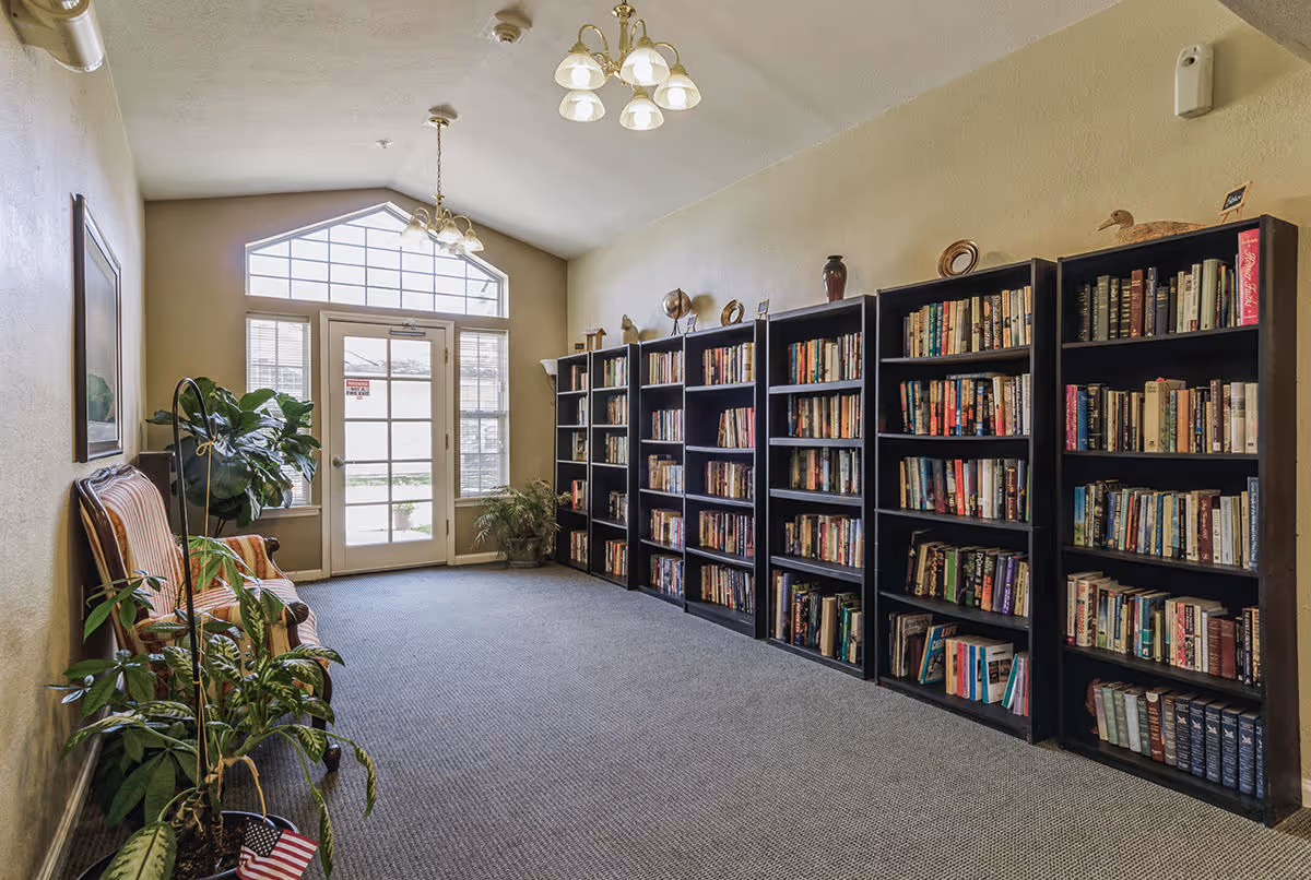 A quiet reading room with several tall black bookshelves filled with books along the right wall. There is a large window and glass door at the far end letting in natural light. On the left side, there is a striped upholstered bench and several potted plants. The room has beige walls, a carpeted floor, and two ceiling light fixtures.